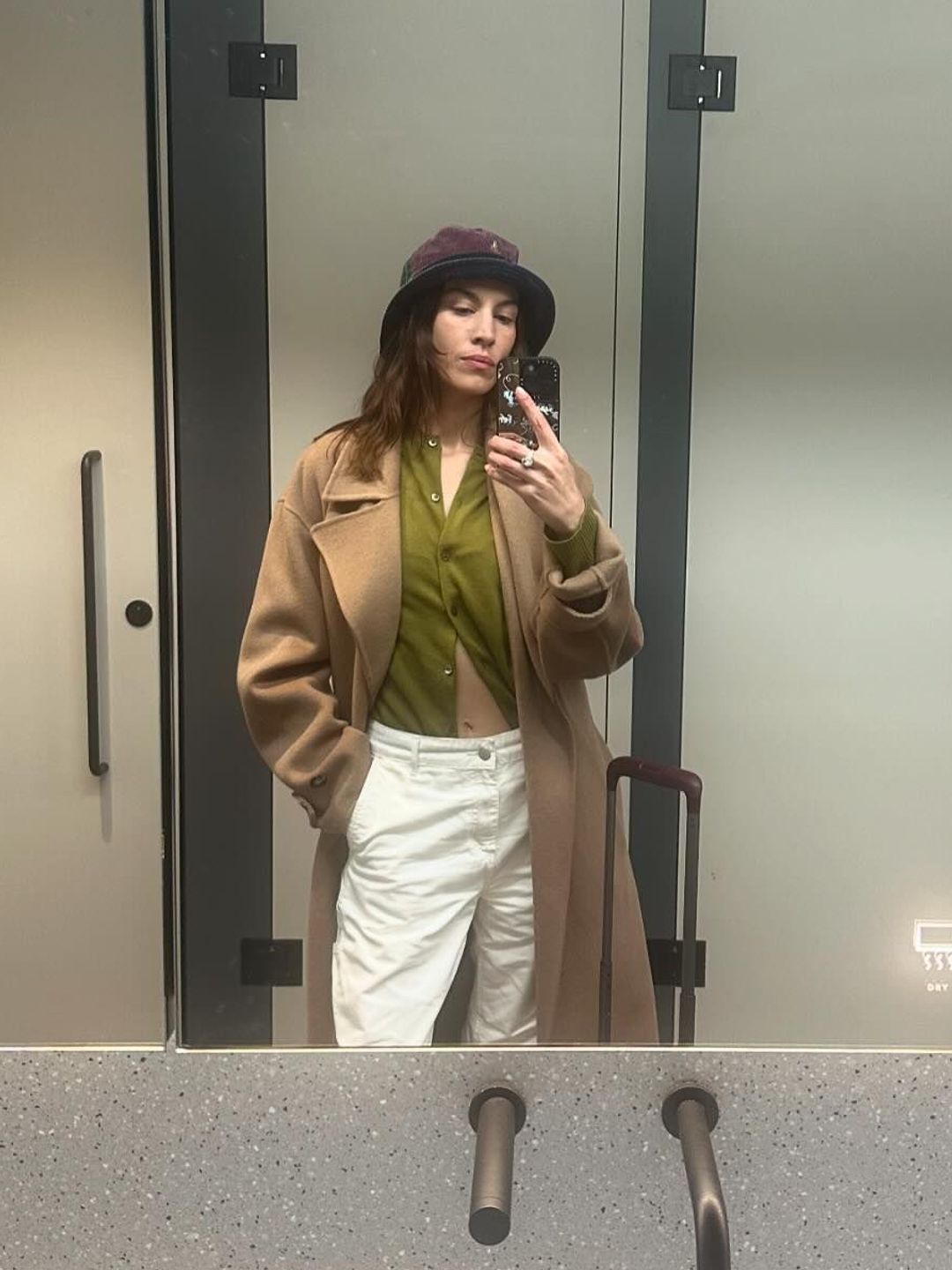 Alexa Chung takes a mirror selfie in white pants, a trench coat and green knit cardigan