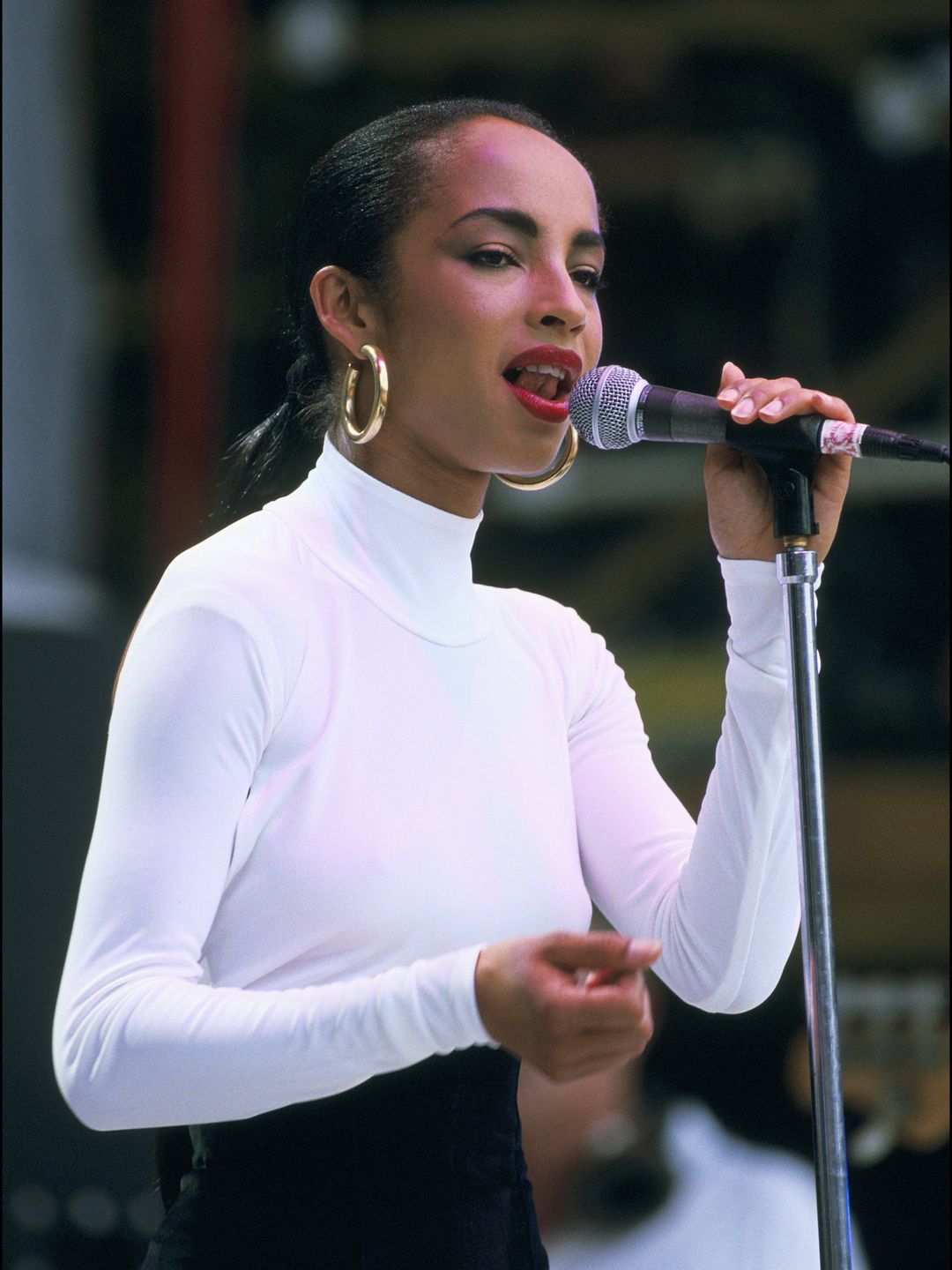 Sade singing in a white top with gold hoop earrings and red lipstick 