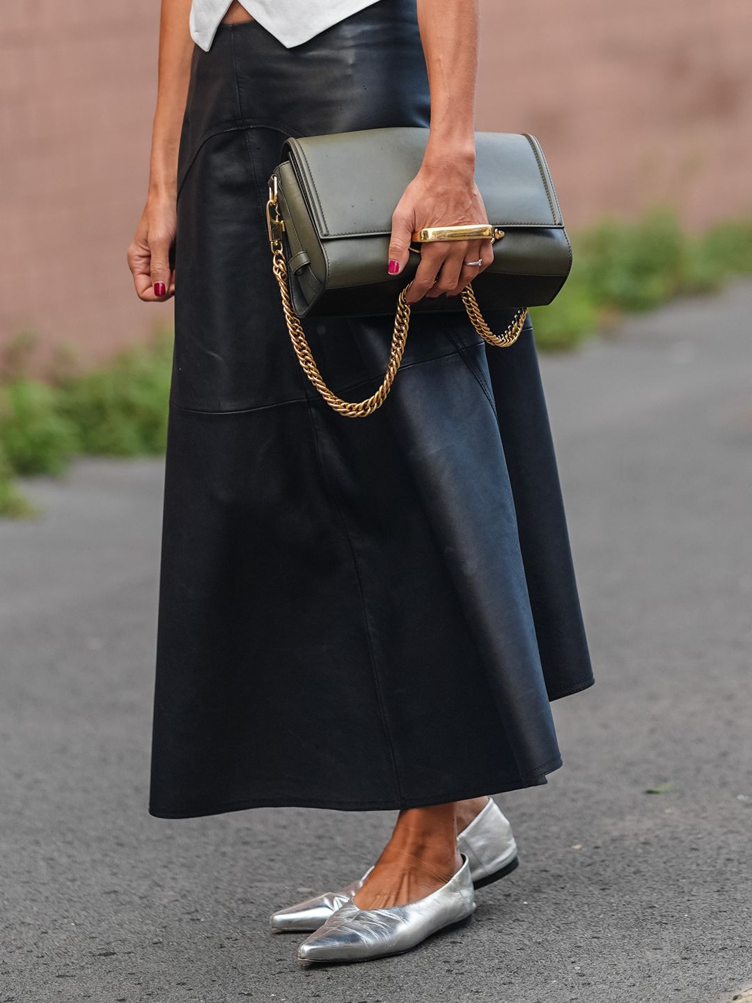 A guest wears black sunglasses, silver earrings, gold necklace, white tailored vest, shiny black leather midi skirt, silver chrome ballerina flats leather shoes, dark green leather bag with gold handle, outside Tod's, during the Milan Fashion Week Spring/Summer 2024-2025 on September 20, 2024 in Milan, Italy. (Photo by Edward Berthelot/Getty Images)