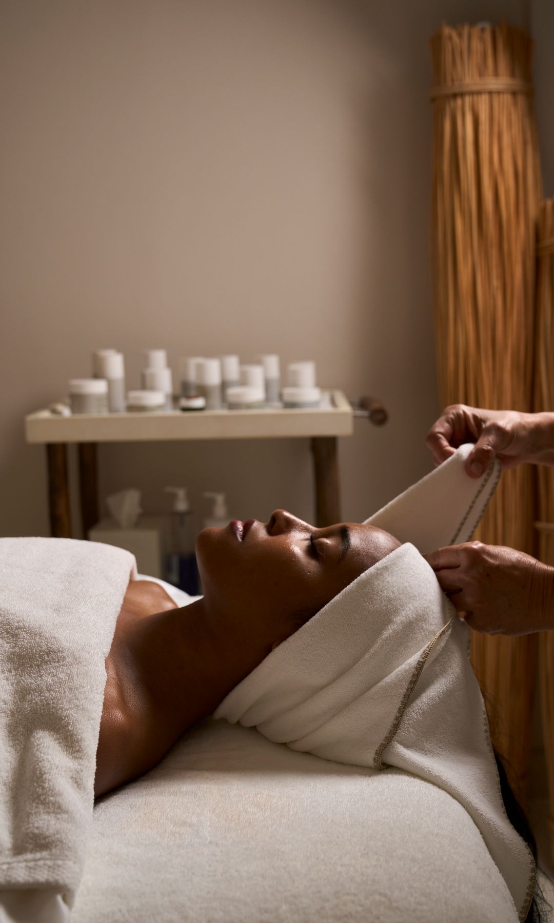 A woman lies on a spa treatment bed with her eyes closed, wrapped in a white towel. A therapist is gently adjusting the towel around her head