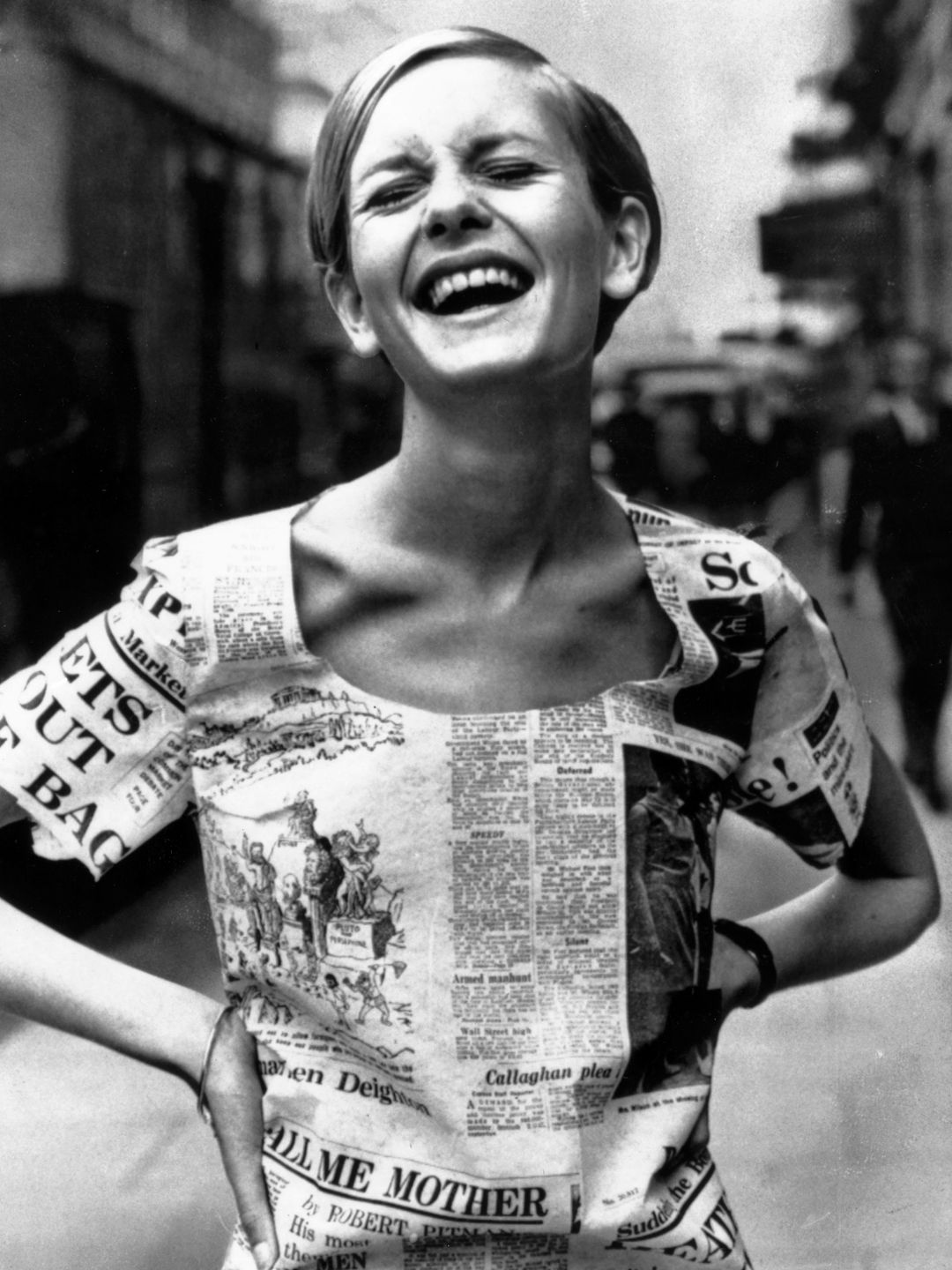 Twiggy incorpora o espírito mod em um vestido estampado de jornal lúdico. Seu sorriso largo e atitude despreocupada representam a exuberância juvenil da moda dos anos 1960. A maquiagem minimalista e o corte de cabelo pixie complementam o vestido ousado, que captura a abordagem vanguardista da era para estampas e padrões.