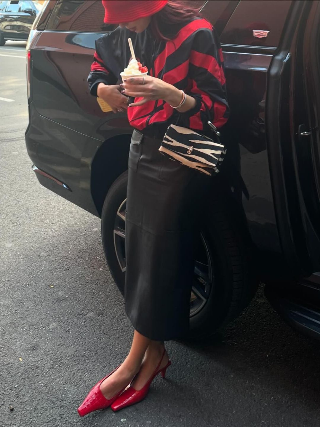 Dua Lipa poses next to a car in a leather midi skirt, red heels and a red and black jacket