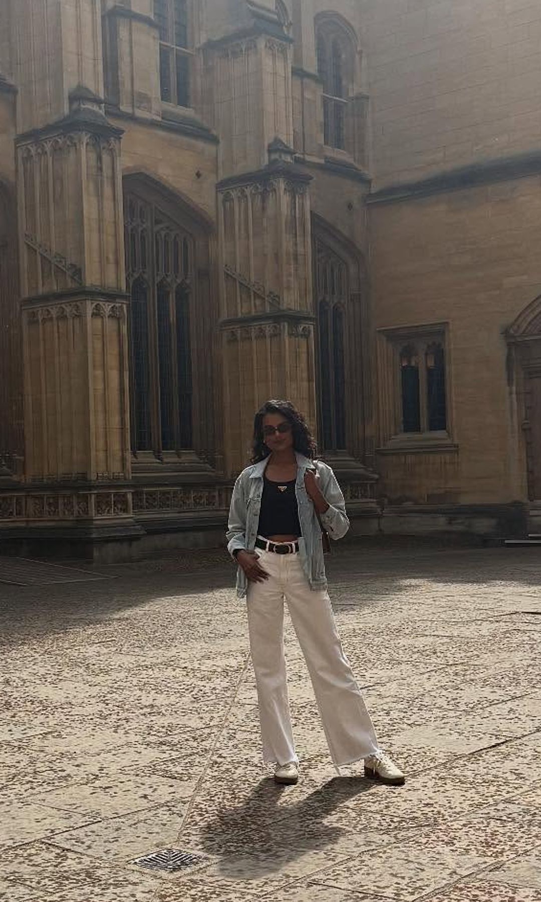 Simone Ashley poses outside of Oxford University in white jeans, sneakers, a black tank top and denim jacket