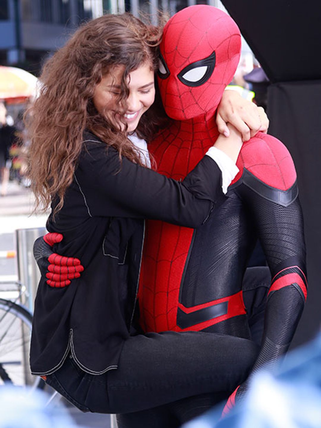 Zendaya holding on to a Spider-Man suited Tom Holland, smiling on set