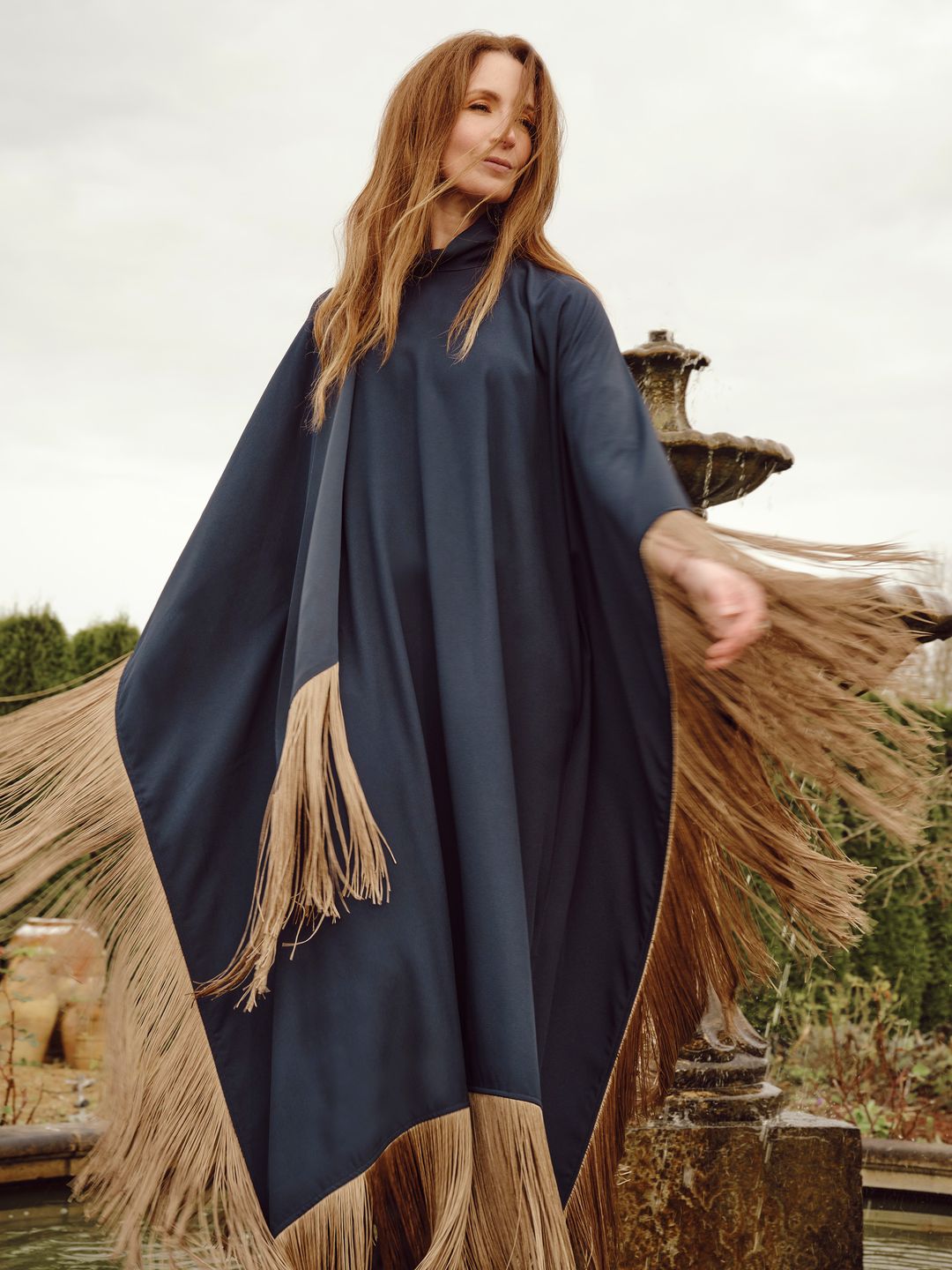 A woman dressed in a navy blue flowing garment adorned with beige fringe detailing stands outdoors in front of a fountain. Her relaxed pose and gentle smile enhance the serene atmosphere.