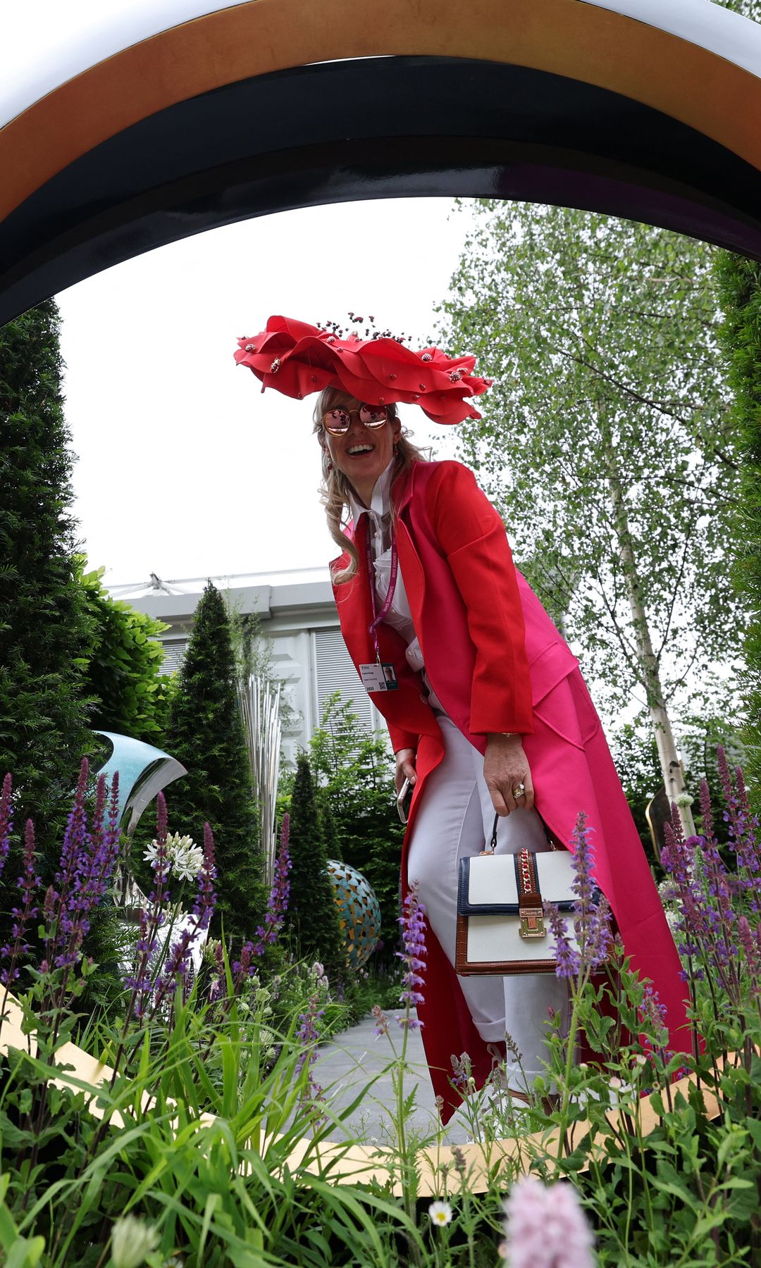 Fellow of the RHS, Laura Konig in a red poppy fascinator and hot pink coat poses through a golden garden sculpture at Chelsea Flower Show.