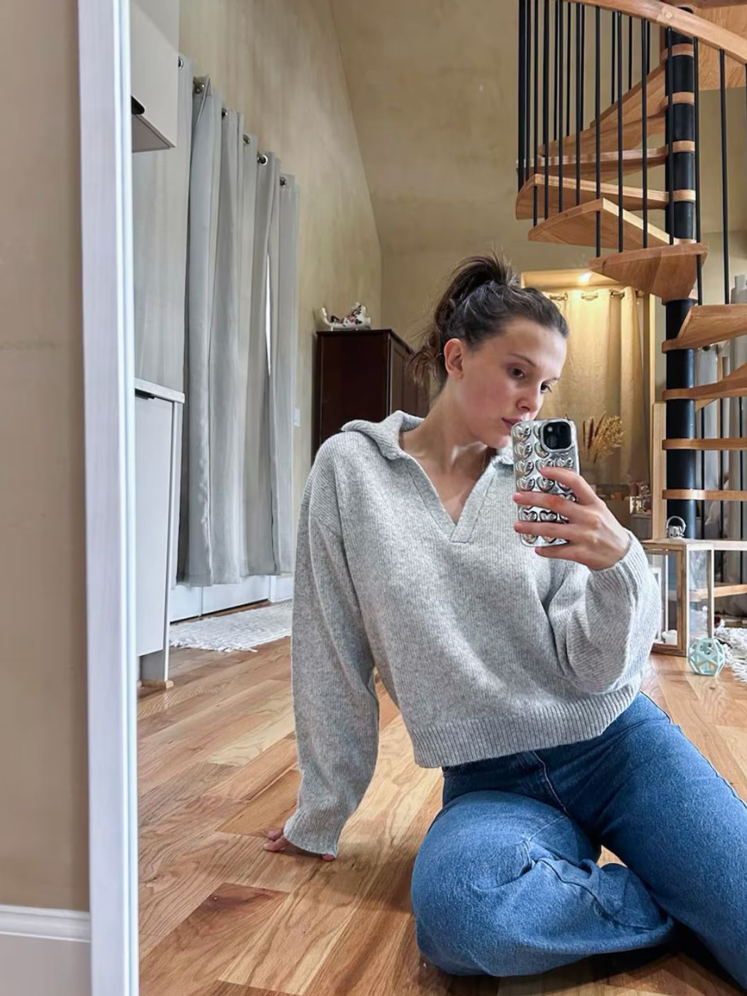 Millie Bobby Brown poses for a mirror selfie in a grey knit and blue jeans