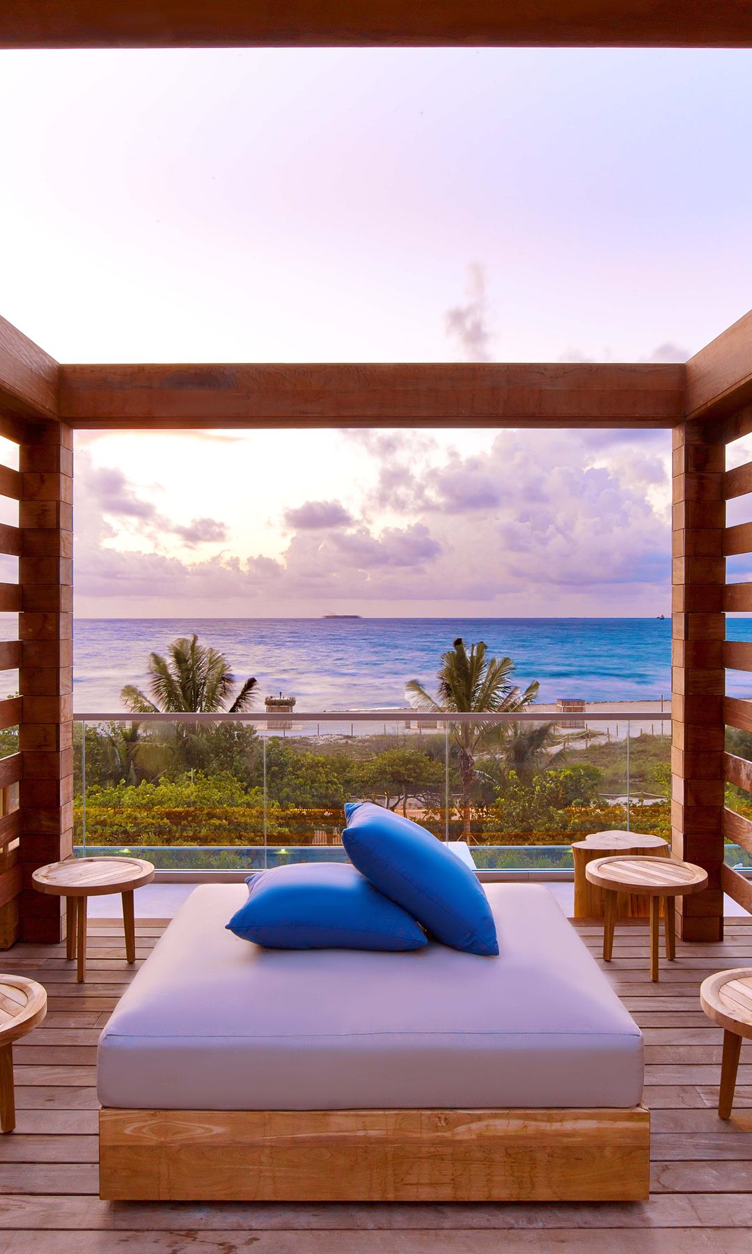 A private wooden cabana at the 1 Hotel South Beach pool, featuring a cushioned daybed with blue pillows and surrounded by small round wooden side tables.
