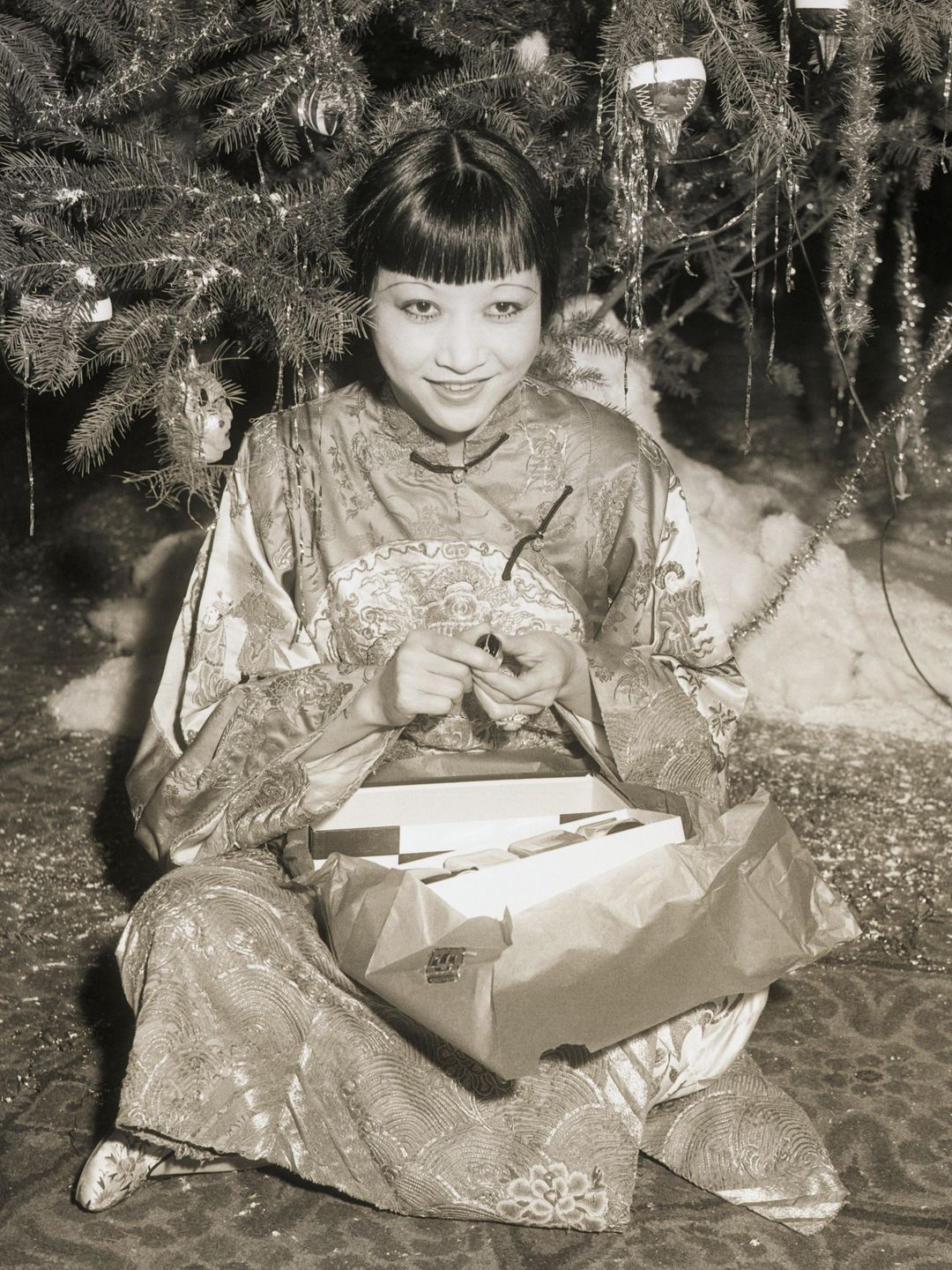 The image is a vintage black-and-white photograph. It features a woman sitting on the floor under a decorated Christmas tree. She is dressed in an ornate traditional outfit, resembling a silk Chinese gown with intricate embroidery. She is smiling warmly and appears to be holding an item, possibly a gift, in her hands. There is wrapping paper and a box near her, suggesting she has just opened a present. The floor is carpeted with a patterned design, and the Christmas tree is adorned with tinsel, ornaments, and lights. The setting exudes a festive and nostalgic atmosphere.