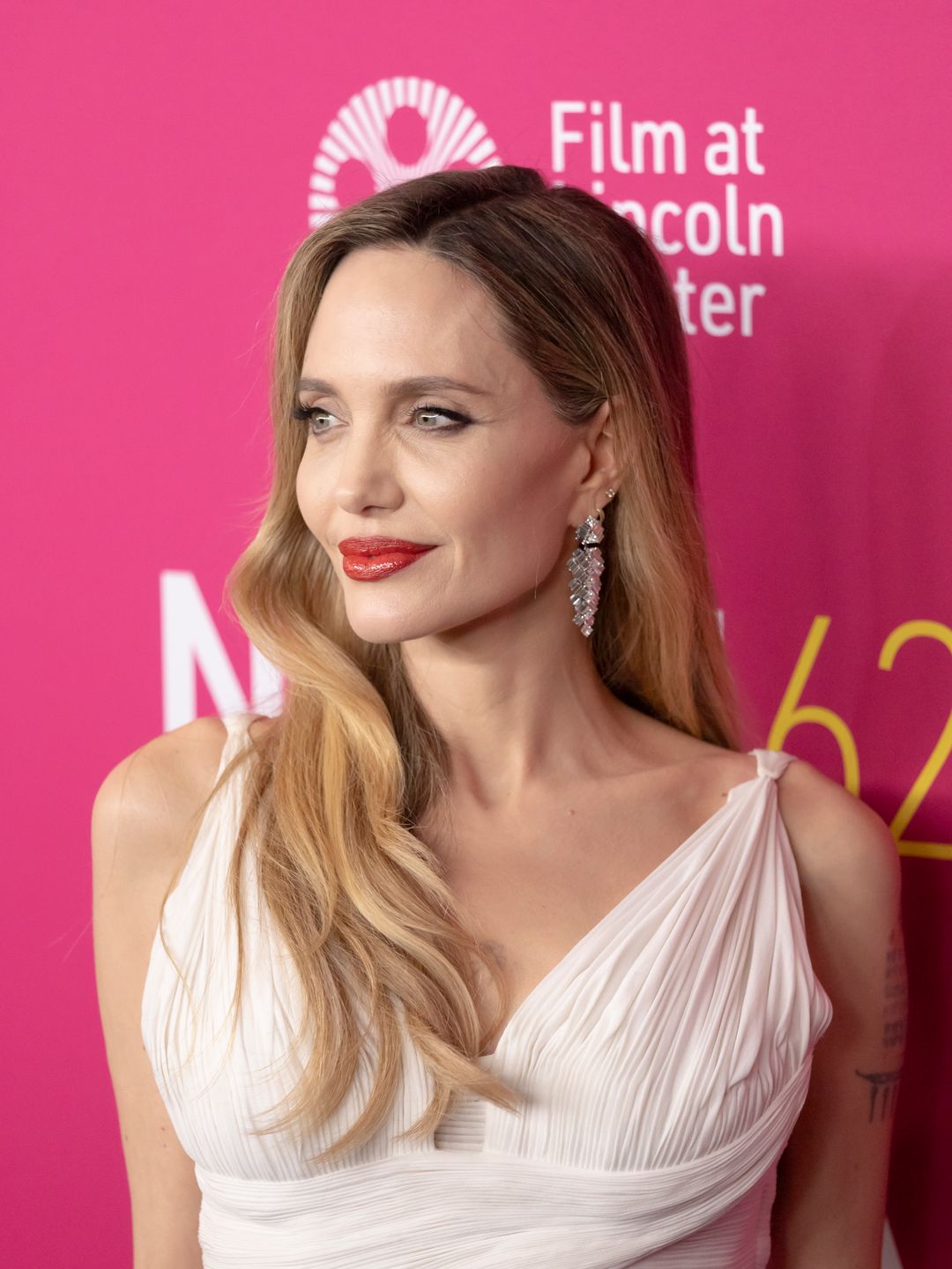 A close-up of Angelina Jolie with blonde, wavy hair and striking red lipstick. She is wearing a white, pleated gown with thin straps, posing in front of a pink background with yellow text.