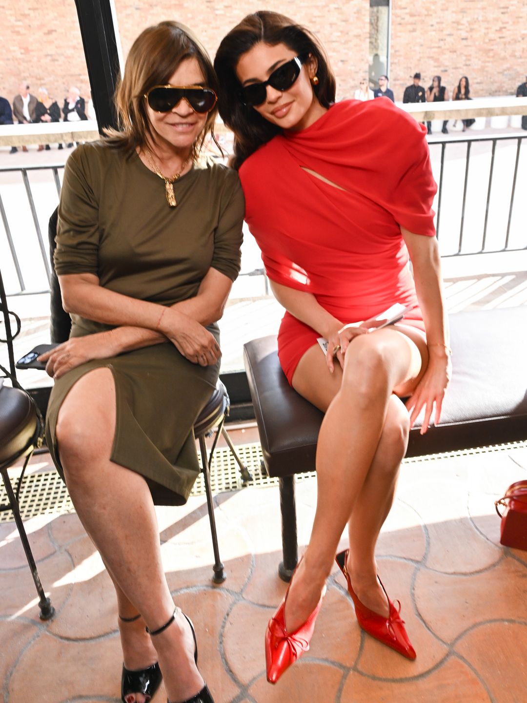 Carine Roitfeld and Kylie Jenner smile for the camera at the Jacquemus Fashion Show. Kylie wears a red dress and matching heels where Carine wears a khaki dress