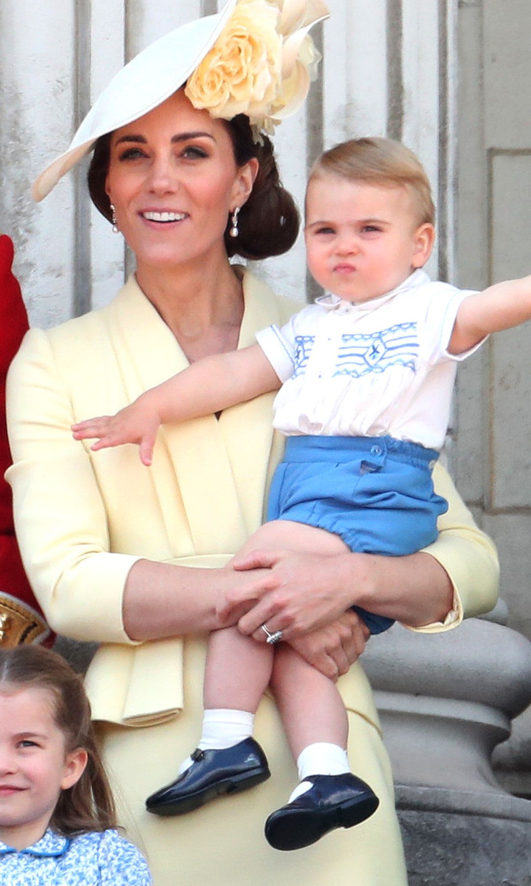 Prince Louis in blue shorts waving while Princess Kate held him