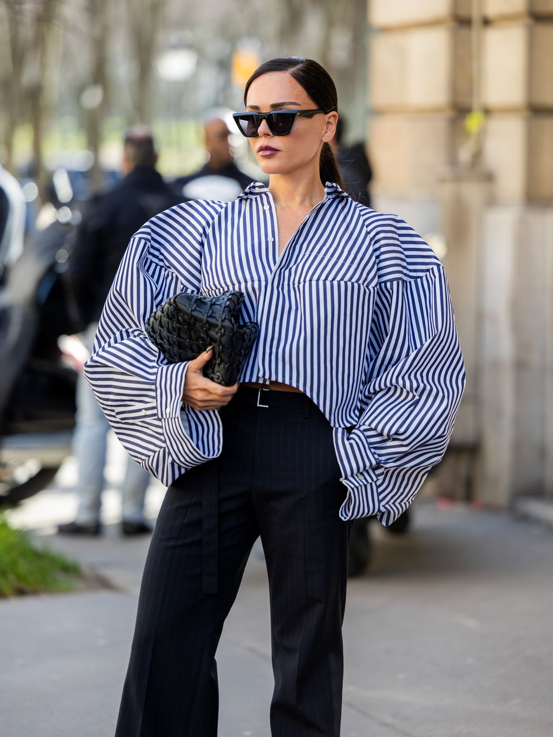 angelie Smyrniotaki wears oversized blue white striped button shirt, black bag, flared high waisted pants outside Sacai during the Womenswear Fall/Winter 2024/2025 as part of  Paris Fashion Week