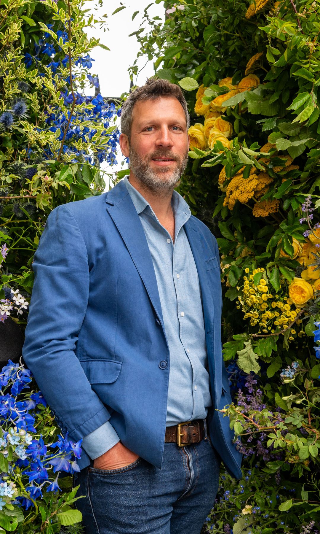 Luxury florist Colin Gray stands in front of a lush wall of flowers wearing a blue blazer and denim, echoing the vibrant floral tones around him.