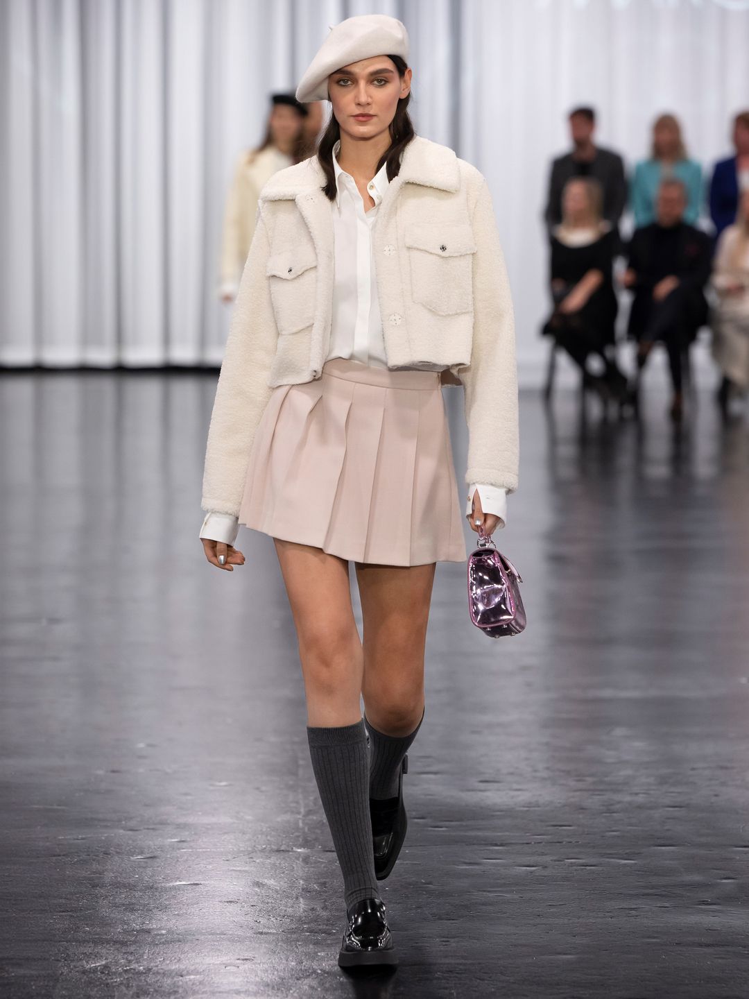 A model at Marc Cain wears a pleated skirt with a white crop jacket, beret, knee high socks and loafers