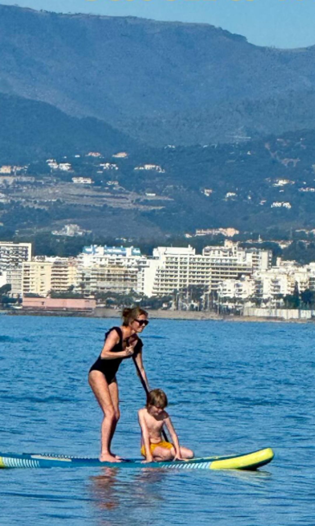 Cat Deeley and her son on a surfboard with a city behind them