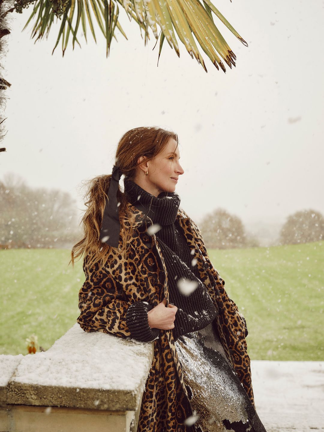 A woman wearing a leopard-print coat and a chunky black turtleneck sweater leans against a stone wall. Snow falls gently around her, with a scenic snowy field and palm fronds in the background.