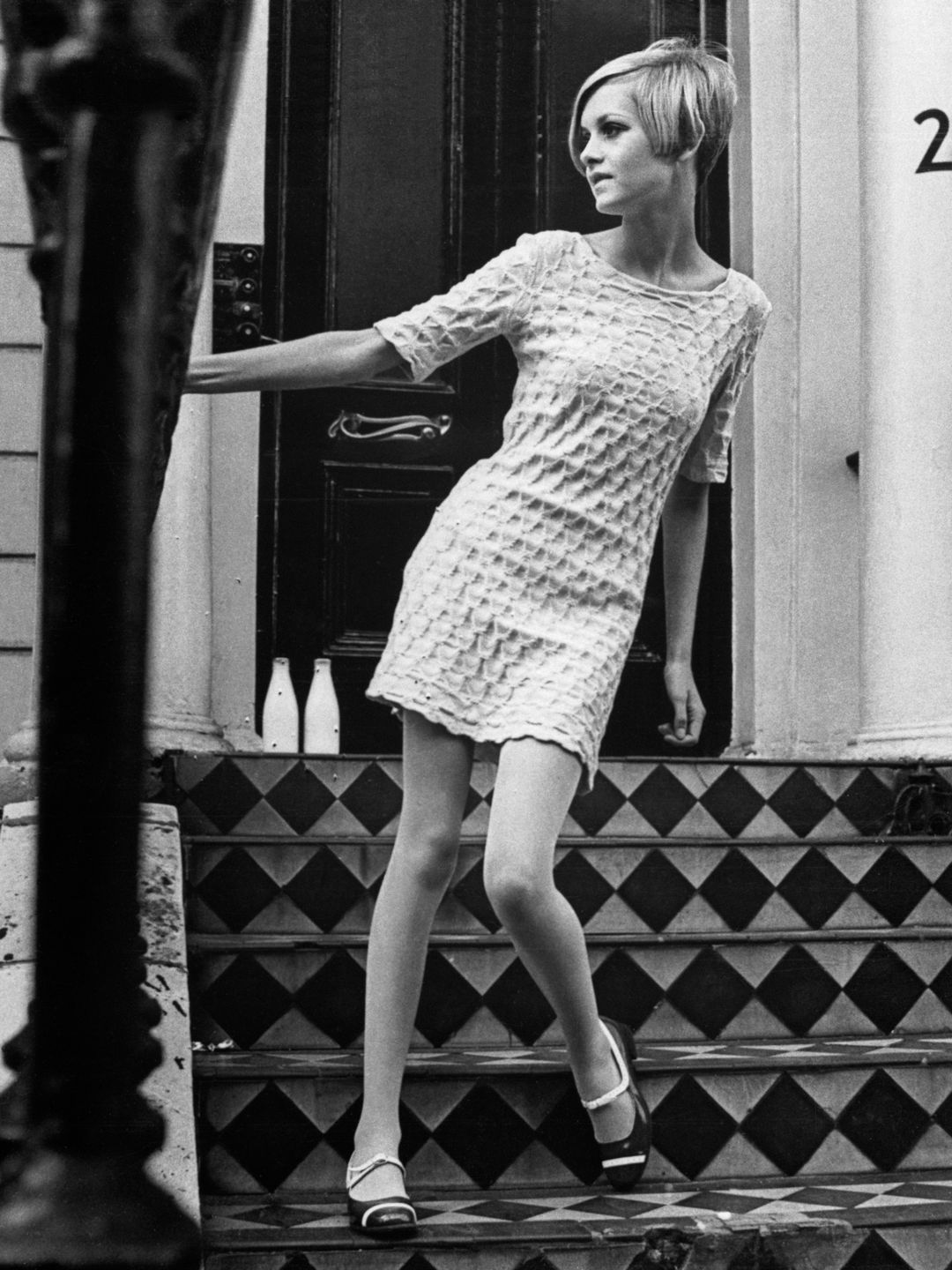 Twiggy pisa confiantemente em uma escada quadriculada em um minivestido branco texturizado. A cena monocromática destaca sua silhueta moderna e estilo minimalista