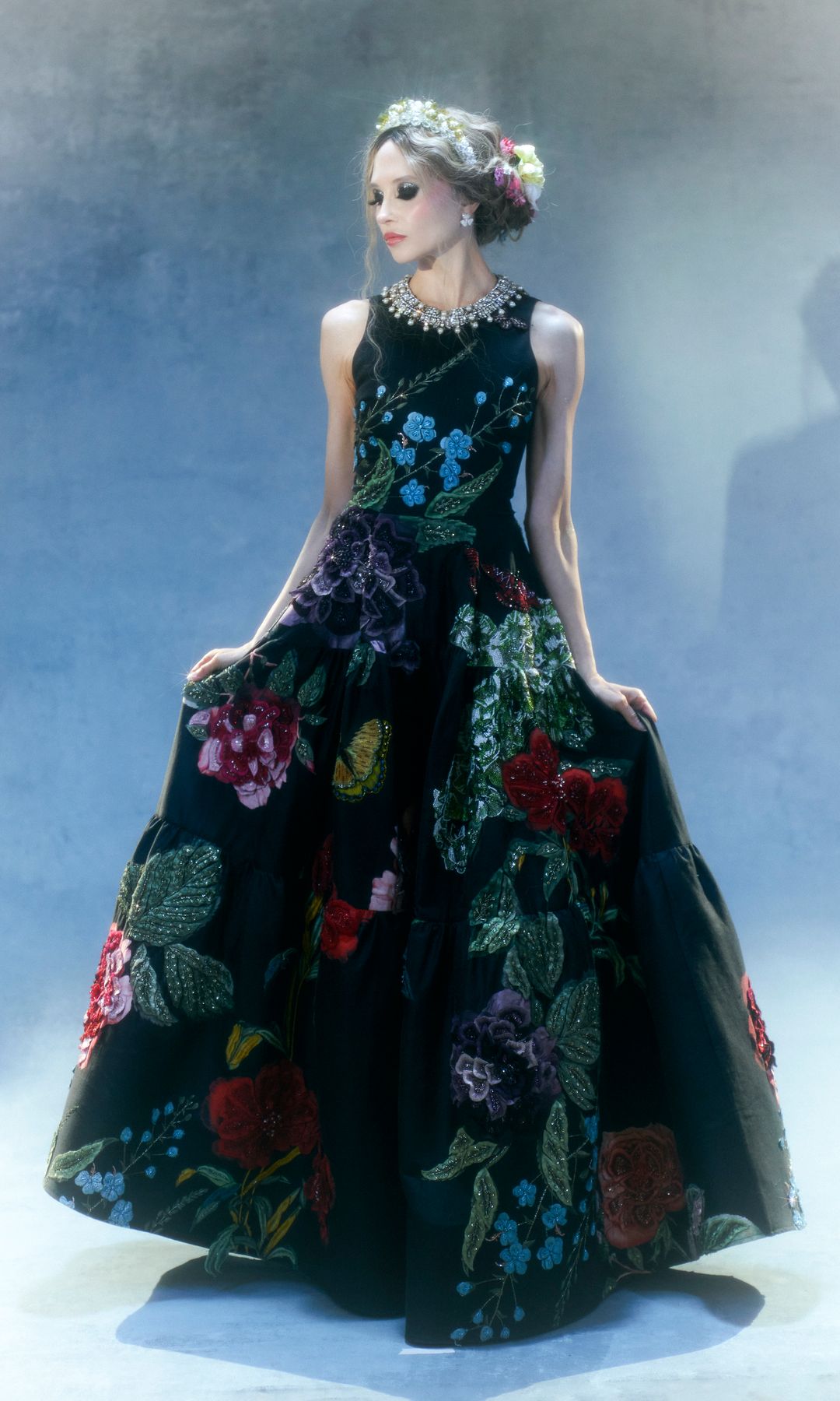 Stacey wears a breathtaking black ballgown covered in vivid floral embroidery. The scene is ethereal, with soft lighting creating a dreamy, misty atmosphere. Her hair is adorned with flowers and her neck sparkles with a statement necklace.