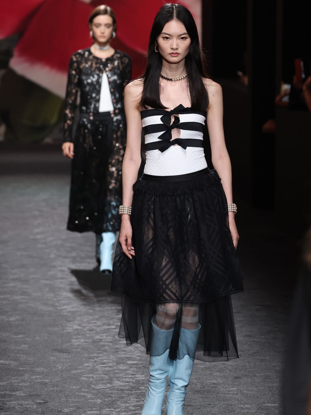 A model walks for the SS24 Chanel show wearing a sheer black chiffon skirt, white and black strapless top, teamed with baby blue knee high boots in a classic Chanel shape 