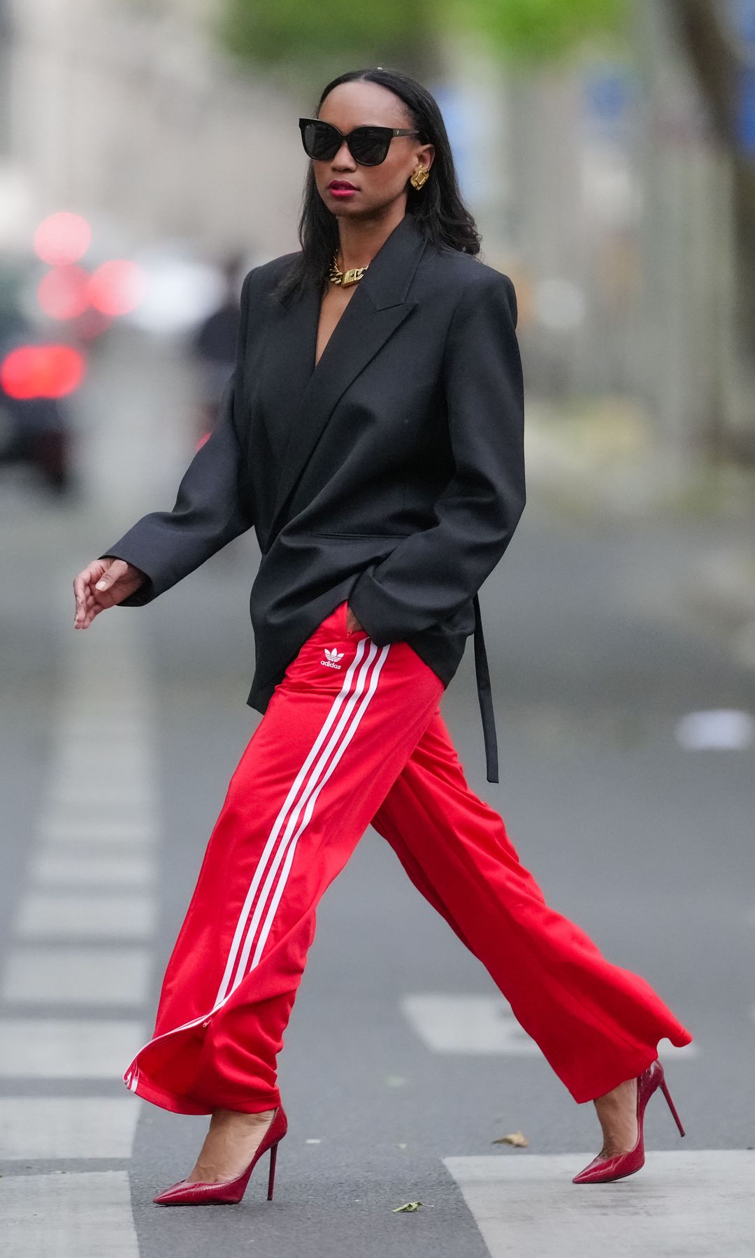Emilie Joseph wears sunglasses, earrings, red lipstick, a black oversized / structured virgin wool blend Blazer with lapel-collar by Victoria Beckham for Mango, Adidas Classics oversized tracksuit bottoms / track pants in red with stripes, a golden chunky necklace / chain by Merbabe Greece, shoes
