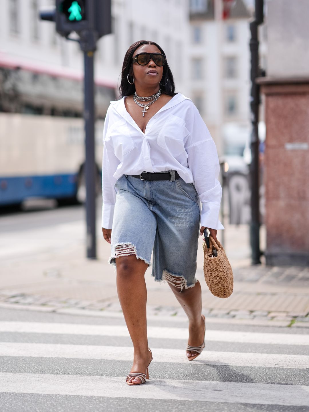 A guest wears black sunglasses, silver hoop earrings, silver layered choker necklaces, white button up long sleeve oversized shirt, black belt, light gray ripped denim jean shorts, light brown woven bag, dark brown heels leather shoes, outside Gestuz at Copenhagen Fashion Week. 