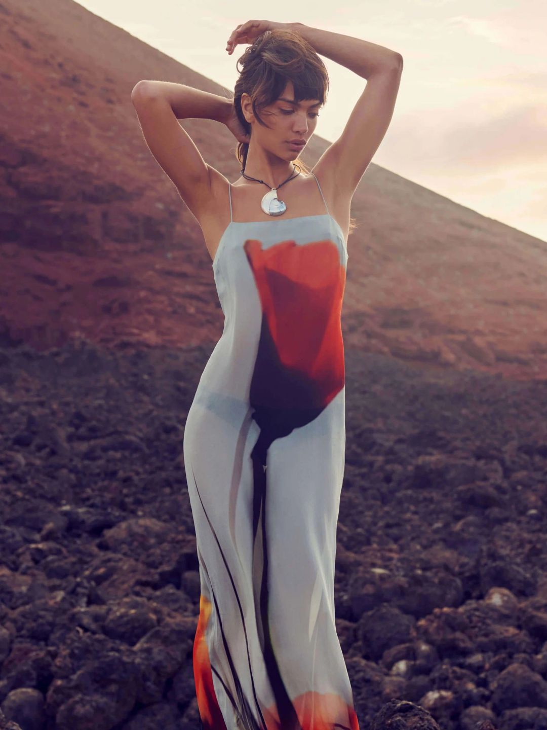 Luciana Maxi Dress - Rat & Boa - This image shows a woman standing against a rugged, rocky landscape with her arms raised, wearing a flowy dress adorned with a large red floral print. Her expression is serene, and the scene feels peaceful and earthy, enhanced by a soft, natural light at dusk. The dress contrasts beautifully with the warm, earthy tones of the background.