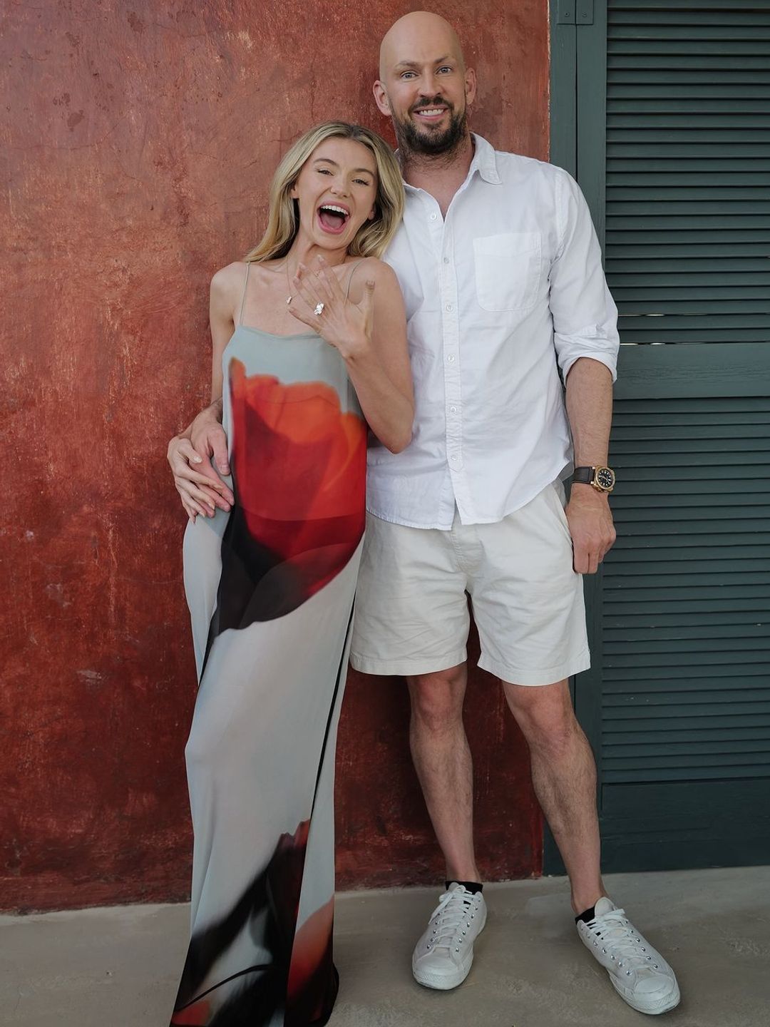 Georgia and James, both smiling, pose side-by-side in a relaxed and happy manner. She flaunts her engagement ring, wearing a flowing floral dress, while he’s dressed casually, against a textured wall backdrop.