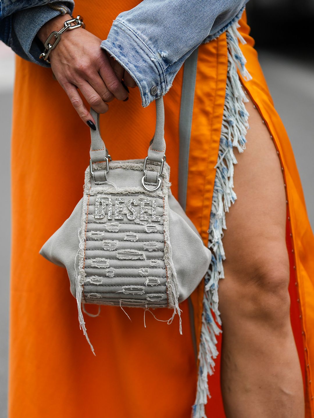 MILAN, ITALY - SEPTEMBER 21: A guest wears silver a pale gray denim ripped jacket from Diesel, an orange long dress with blue denim fringed borders, a beige denim / embroidered rhinestones handbag from Diesel, outside Diesel, during the Milan Fashion Week