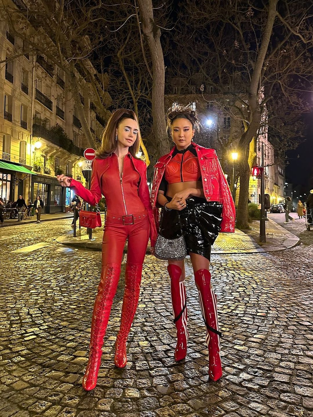 The image features a scene from Emily in Paris, showing actors Lily Collins (left) as Emily and Ashley Park (right) as Mindy. They stand on a cobblestone street at night, dressed in bold red outfits—Collins in a leather catsuit and Park in a glossy jacket and skirt, exuding confidence and style.