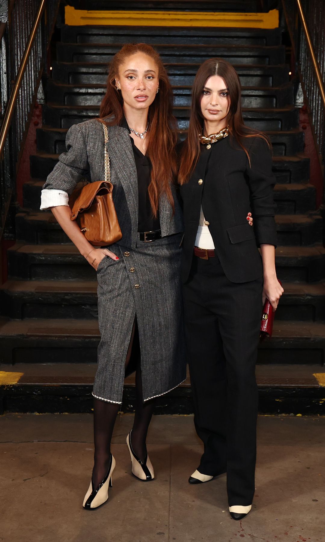 Adwoa Aboah and Emily Ratajkowski pose in the subway. Adwoa wears a grey skirt suit with sheer black tights, monochrome heels and a large brown handbag over her shoulder. Her red hair is worn long and loose.