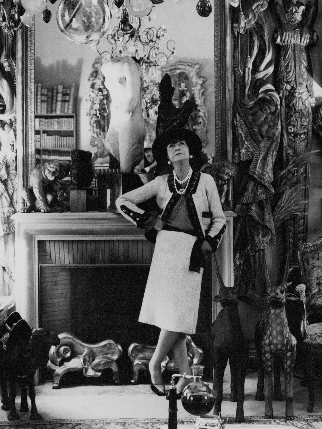 French fashion designer, Coco Chanel, standing, in her Paris apartment, with her back to a mantle fireplace, with a large mirror above, looking up at a chandelier