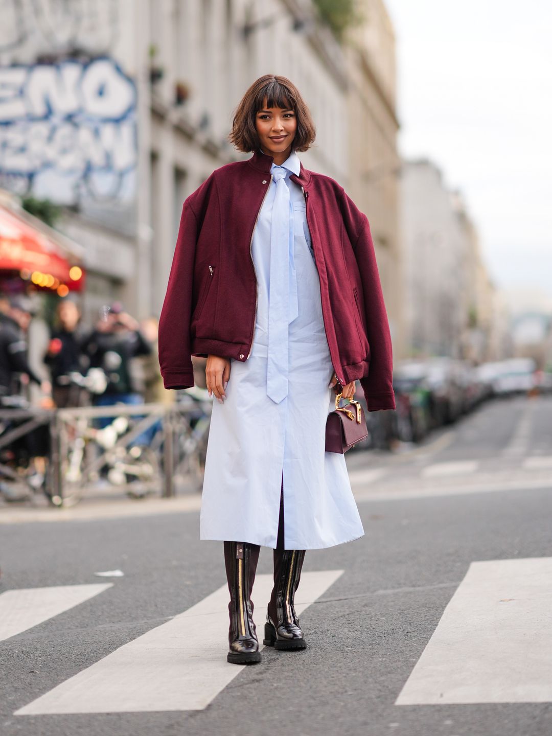 Flora Coquerel wears a burgundy oversized bomber jacket , a pastel pale blue long shirt / midi dress with a tie, a leather bag, knee-high leather boots, outside Casablanca