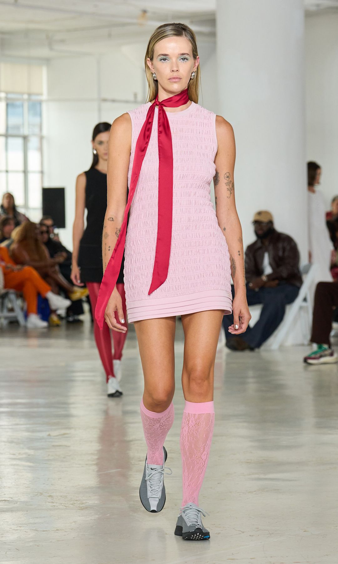 A model wears a pink mini skirt, pink knee high socks and trainers and a red silk scarf tied around her neck