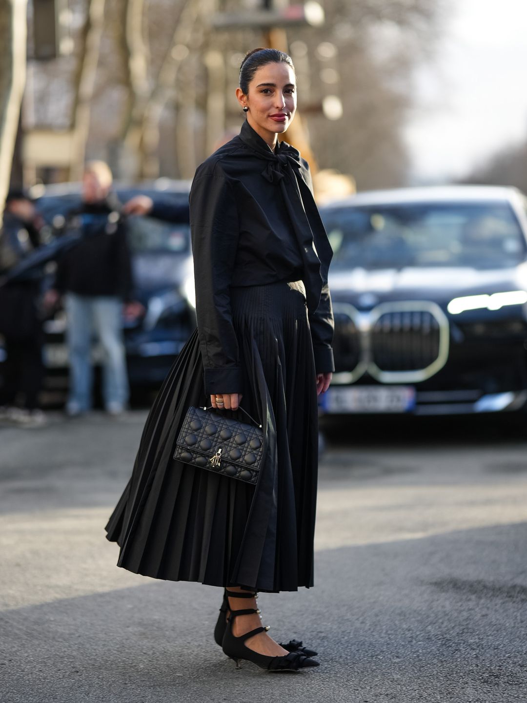 Bettina Looney wears earrings, a black shirt, black tie, pleated midi skirt, quilted leather bag, pointed kitten heels pointed pumps / shoes with bow detail, outside Dior, during the Haute Couture Spring/Summer 2024