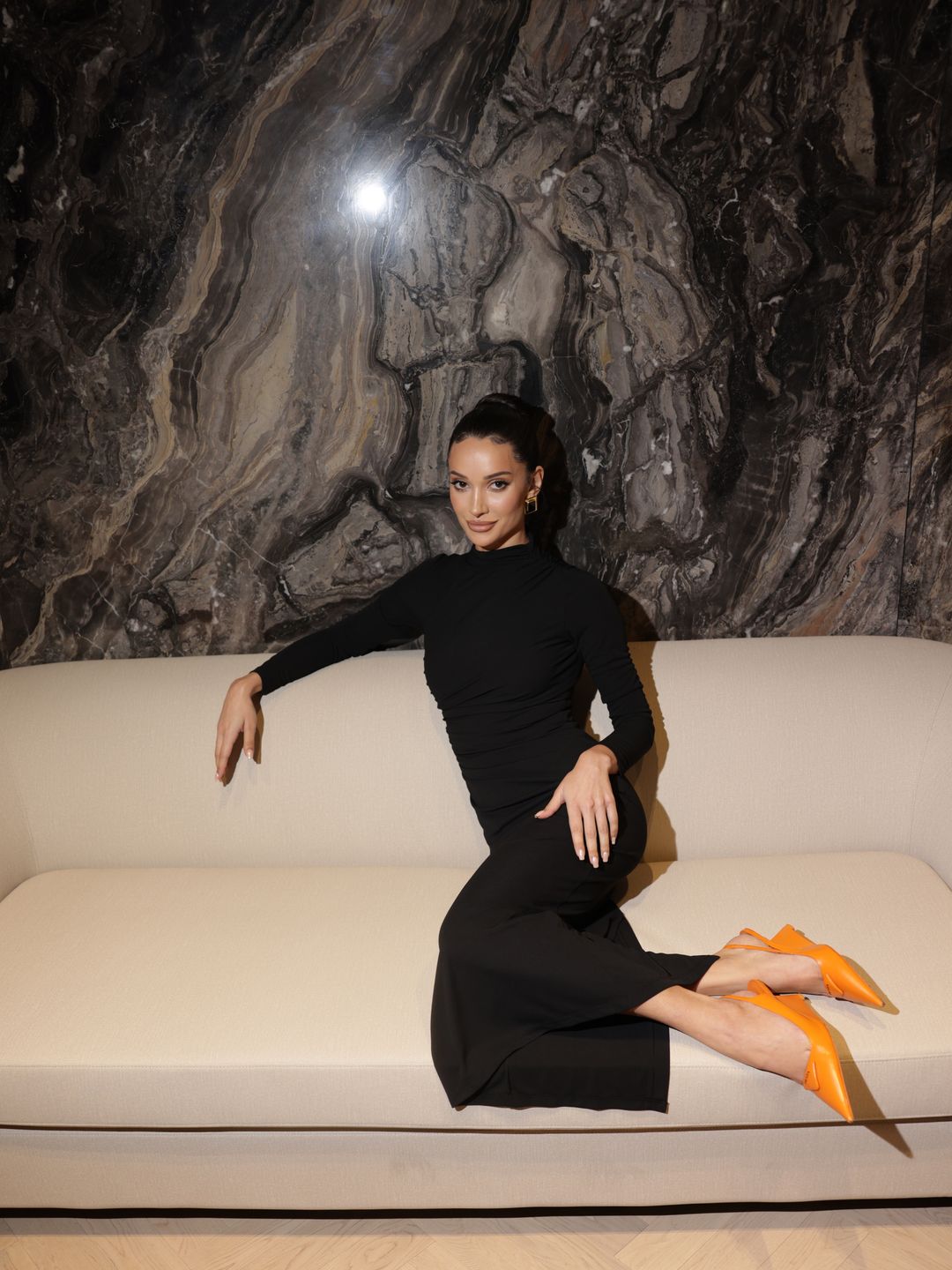 Roxie Nafousi sits elegantly on a cream sofa, leaning slightly to one side. She wears a sleek black, long-sleeved dress paired with bright orange pointed shoes, which stand out boldly. The background features a textured, marble-like wall, creating a dramatic, luxurious ambiance.