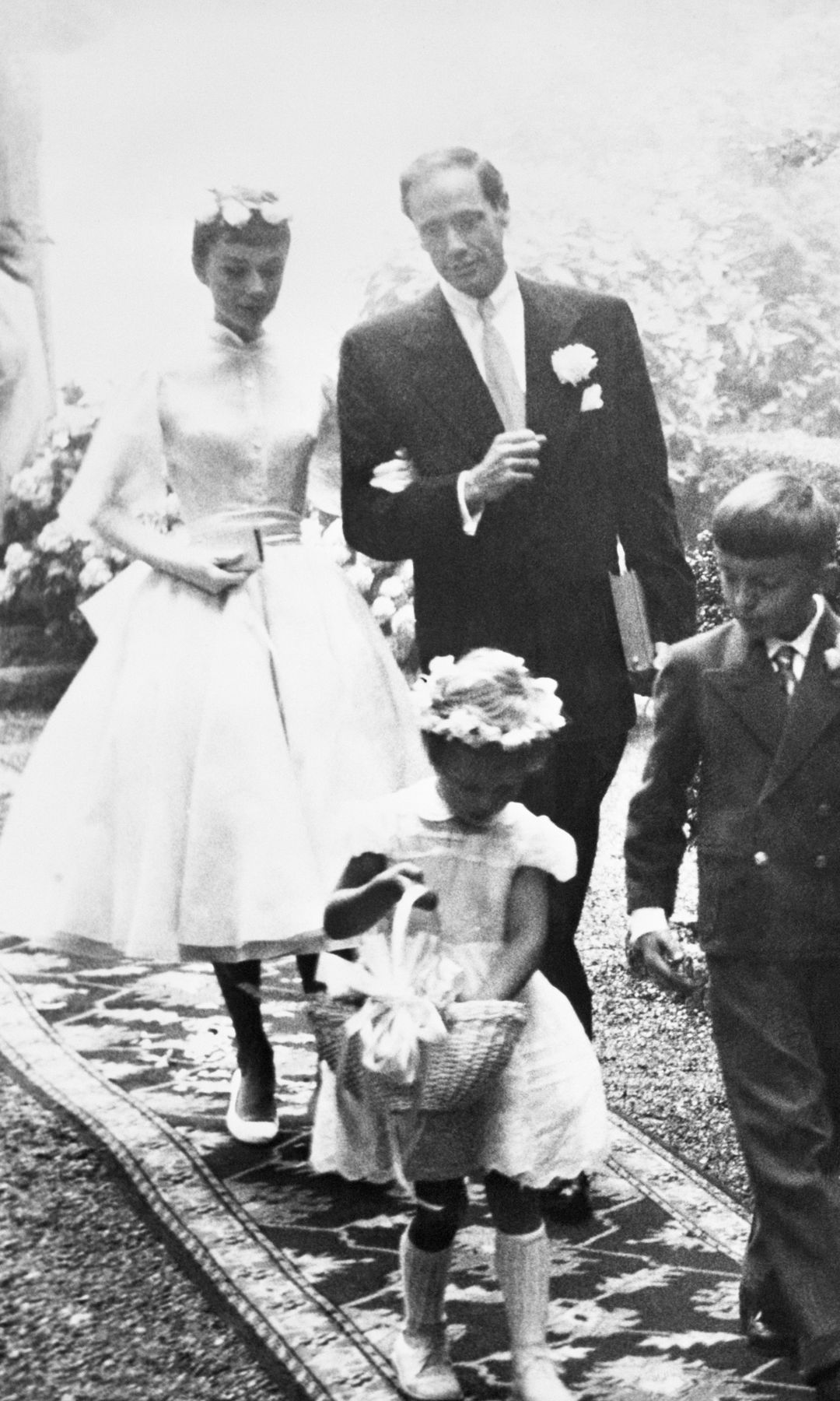 Audrey Hepburn, 25, and Mel Ferrer, 37, leave the chapel after their wedding at Burgenstock Mountain, overlooking Lake Lucerne. The flower girl and page are the children of hotel owner Fritz Frey, whose resort has been the actress's retreat for about ten weeks. Ferrer flew in from Italy for the wedding, which was attended by the Bride's mother, the Dutch Baroness Ella Van Heemstra. A brief secret civil ceremony at Buochs, Switzerland preceded the ceremony.