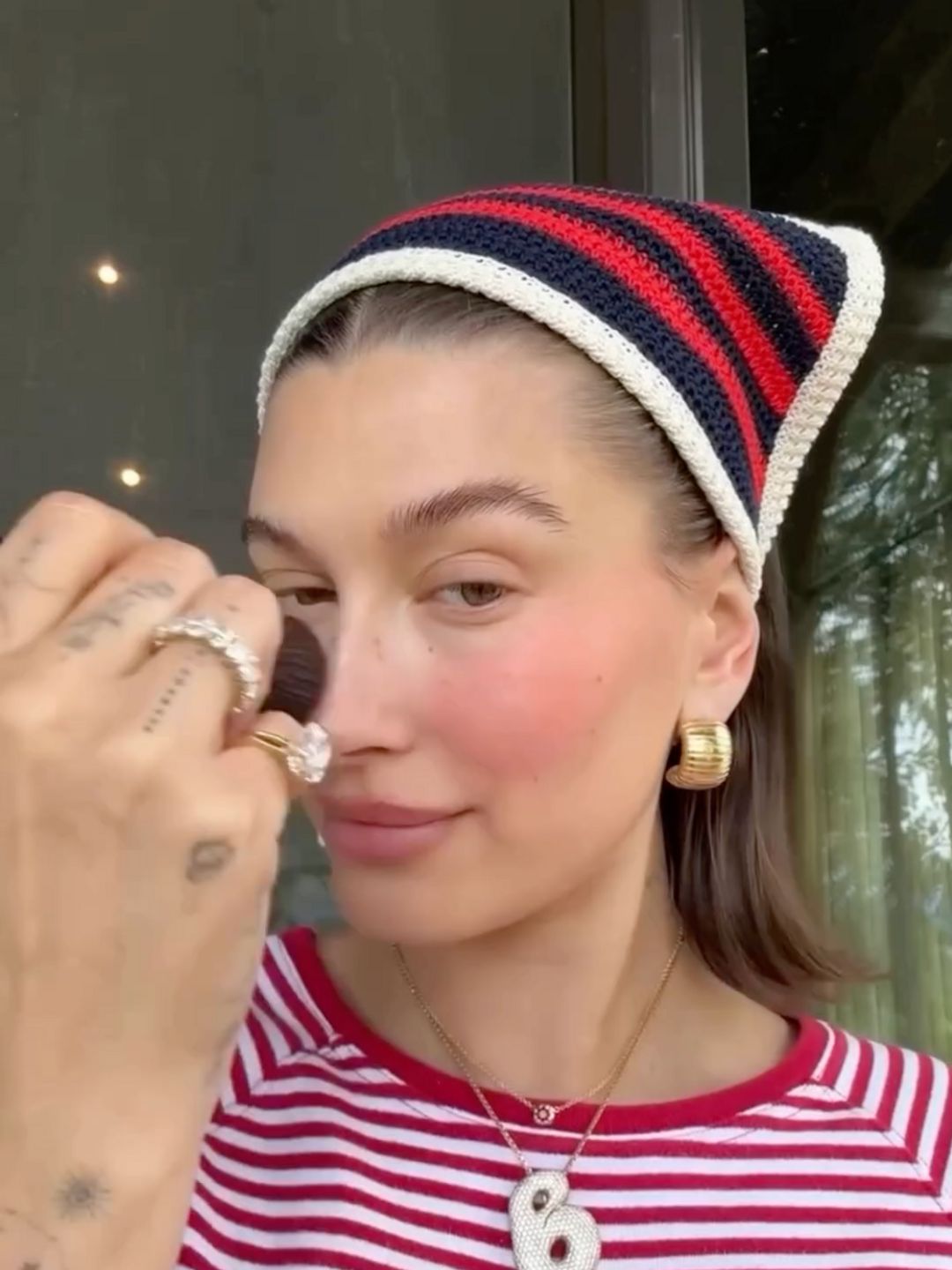 Hailey Bieber does her make up wearing a stripe t-shirt and matching crochet bandana