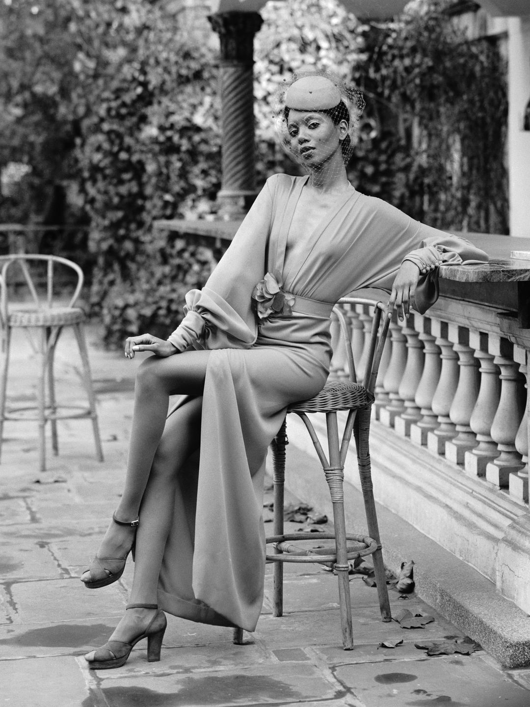 Model sat on a chair wearing a floaty crepe dress and a pillbox hat 