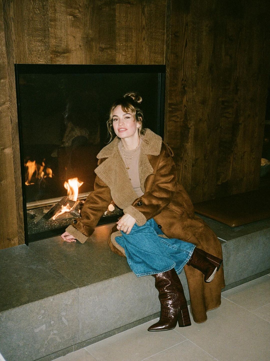 Lily James poses next to a fireplace in jeans and a brown coat