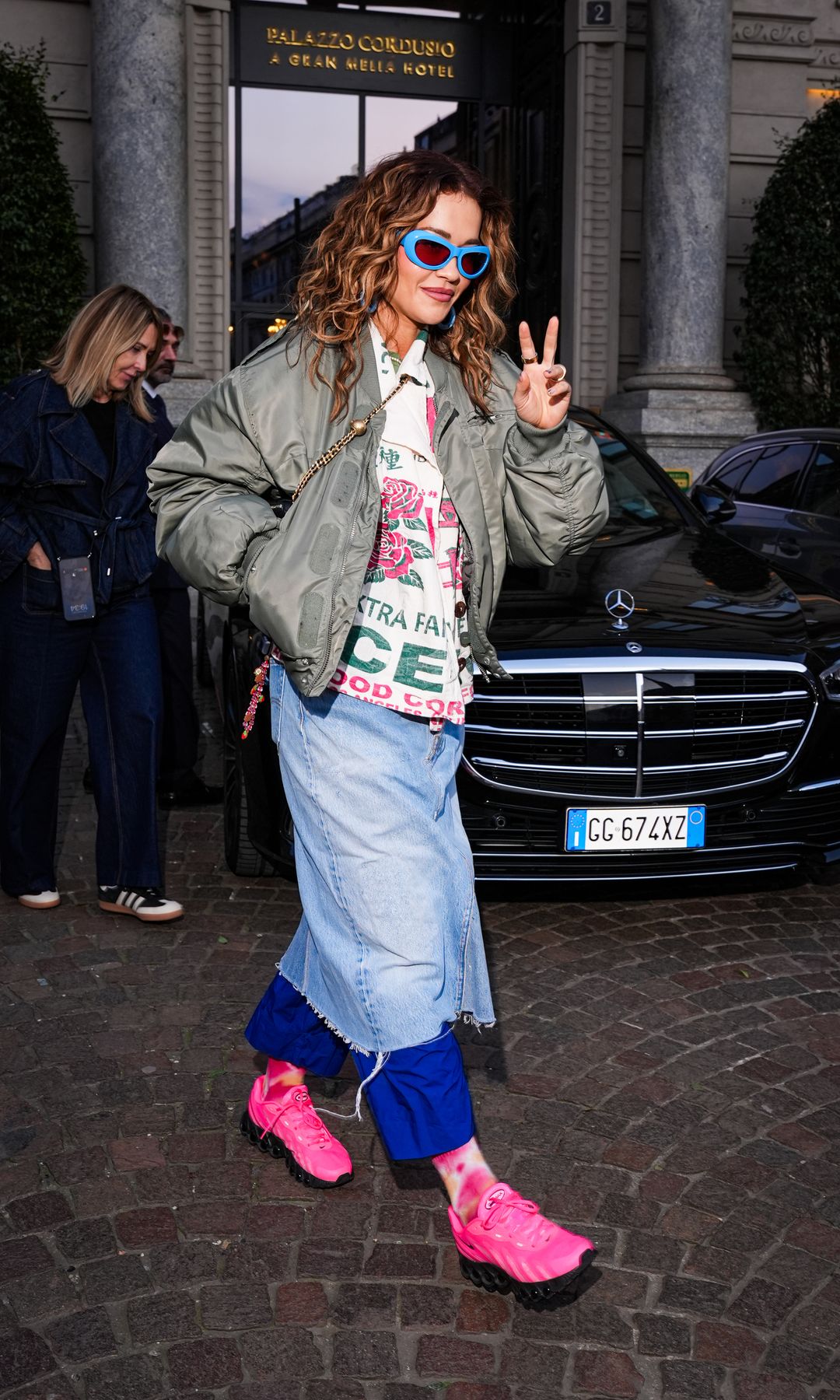 Rita Ora is seen wearing a denim skirt over blue torusers, a button-up graphic shirt and green bomber jacket while outside her hotel in Milan, Italy
