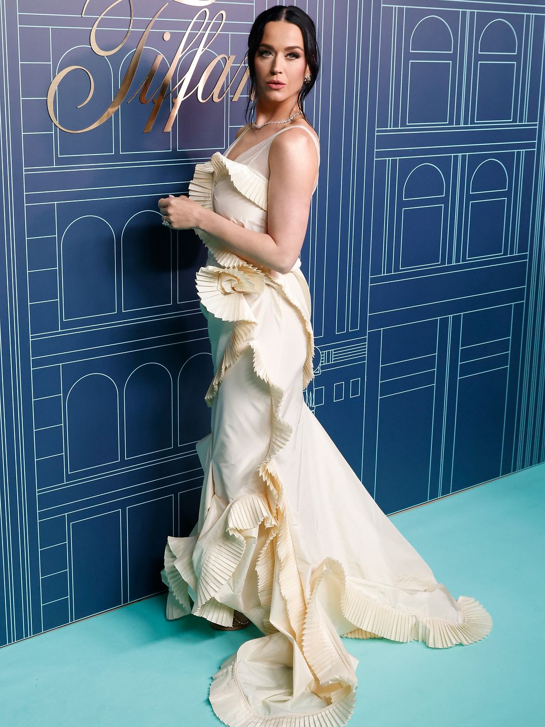 Katy Perry looking over her shoulder, she is wearing a floor length fishtail ivory pleated gown
