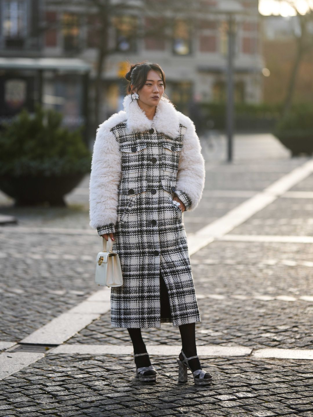 Woman wearing silver large earrings, a black and white checkered tweed pattern with white oversized fluffy sheep collar and sleeves buttoned long coat