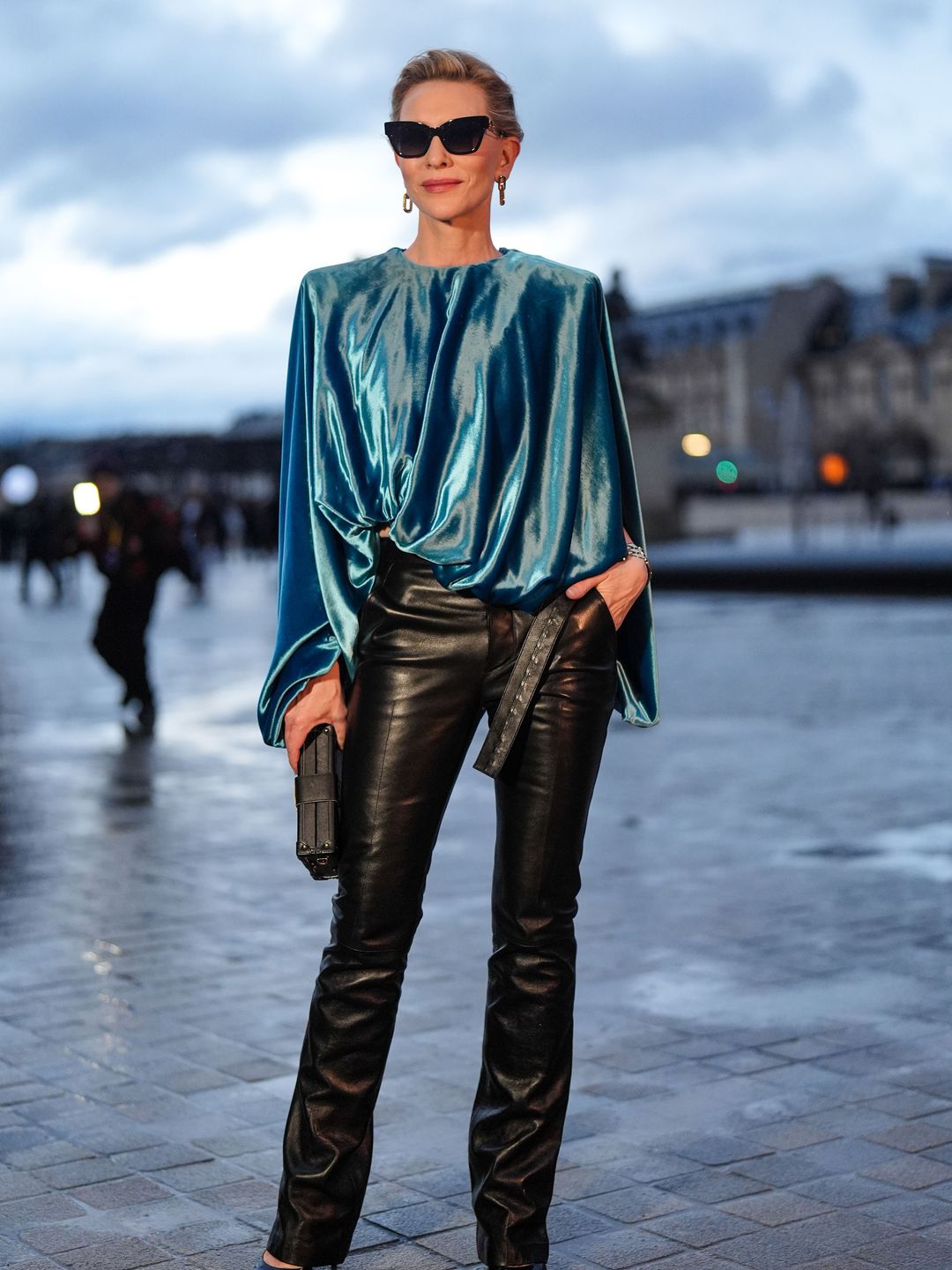 Cate Blanchett wears sunglasses, earrings, a blue turquoise gathered sleeveless lustrous shiny silk top, a bracelet, black leather flared pants and pointed shoes outside the Louis Vuitton show