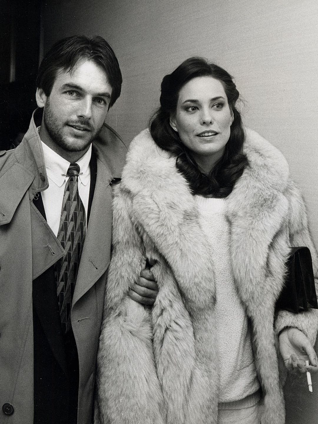 Mark Harmon and Christina Raines at an event. She is wearing a fur coat and he is wearing a trench coat over his suit. 