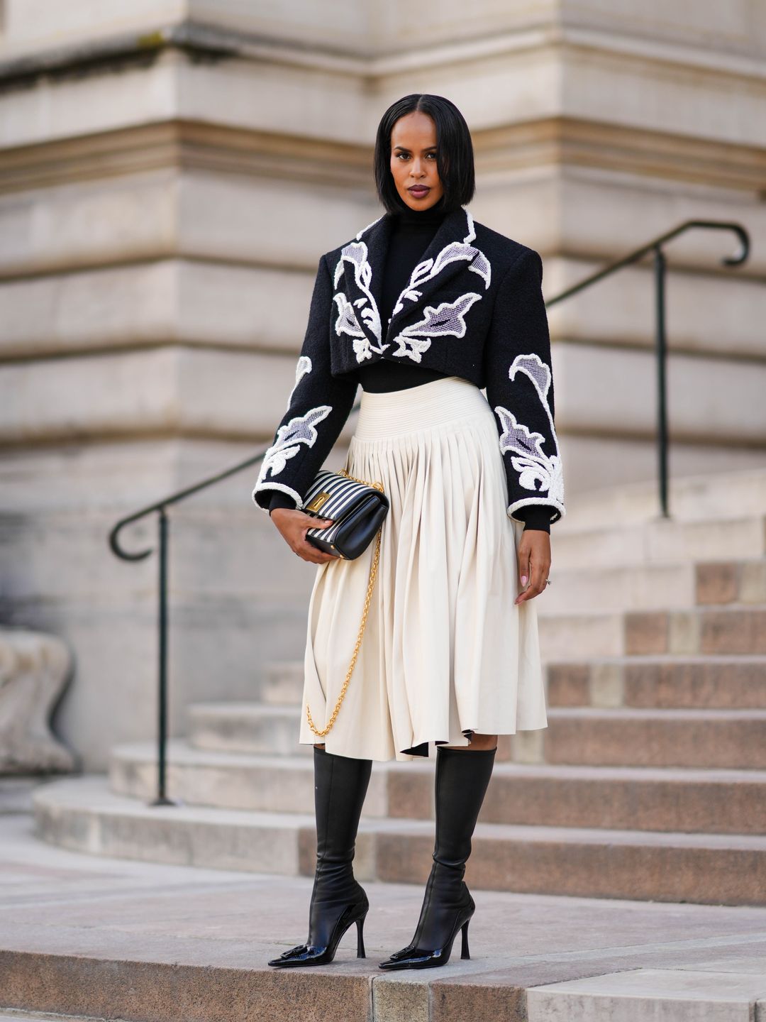 Sabrina Elba wearing a black embroidered jacked with a cream pleated skirt 