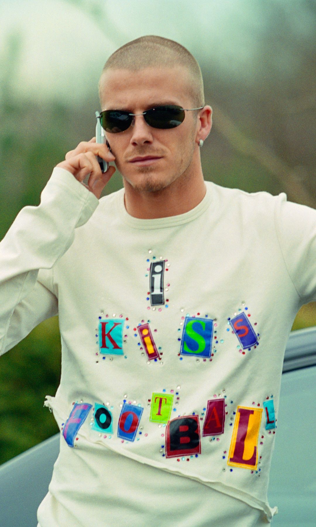 David Beckham leans against a car door while talking on the phone. He is wearing aviator sunglasses and a white long sleeve sweatshirt which has I Kiss Football appliqued onto it. 