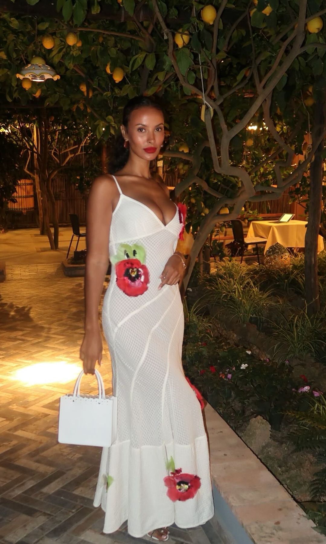 Maya Jama poses under a lush canopy of lemon trees, likely in an al fresco dining setting, possibly in Italy or the South of France. She wears a sheer, white crochet maxi dress that hugs her curves elegantly, adorned with large, vibrant red poppy appliqués. The dress features thin spaghetti straps and a plunging neckline, striking a perfect balance between sultry and sophisticated. She accessorizes with a structured white mini handbag and white sandals, her deep red lipstick echoing the floral embellishments on her dress. Her curly hair is styled to one side, revealing her glowing complexion and gold hoop earrings. She stands on a beautifully tiled floor, with warm ambient lighting adding to the romantic, Mediterranean vibe.