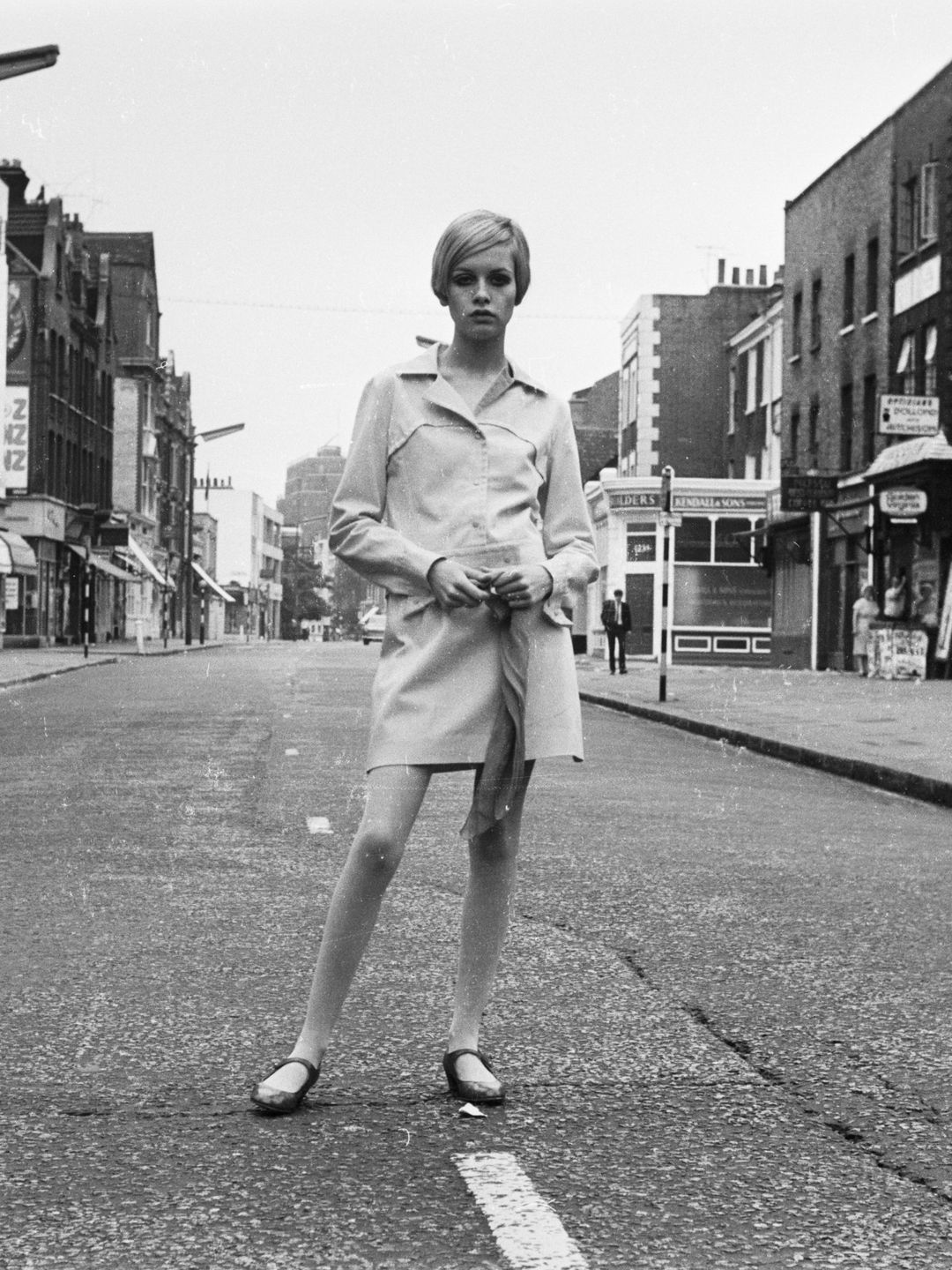 Twiggy está sozinha em uma rua vazia, modelando um mini casaco simples e estruturado que simboliza o estilo mod. Sua pose, juntamente com o cenário urbano austero, destaca seu papel como pioneira na fotografia de moda