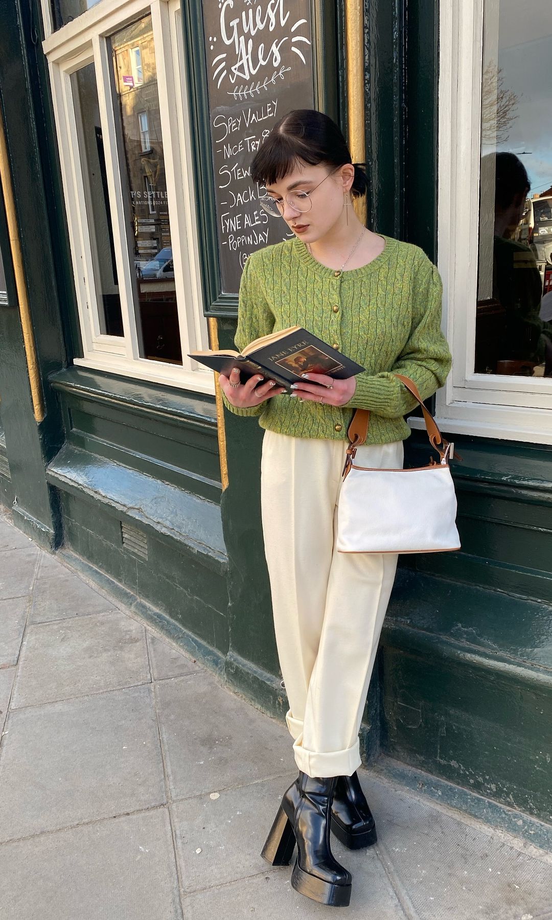 A girl poses in a green knit, cream trousers and boots while reading a book